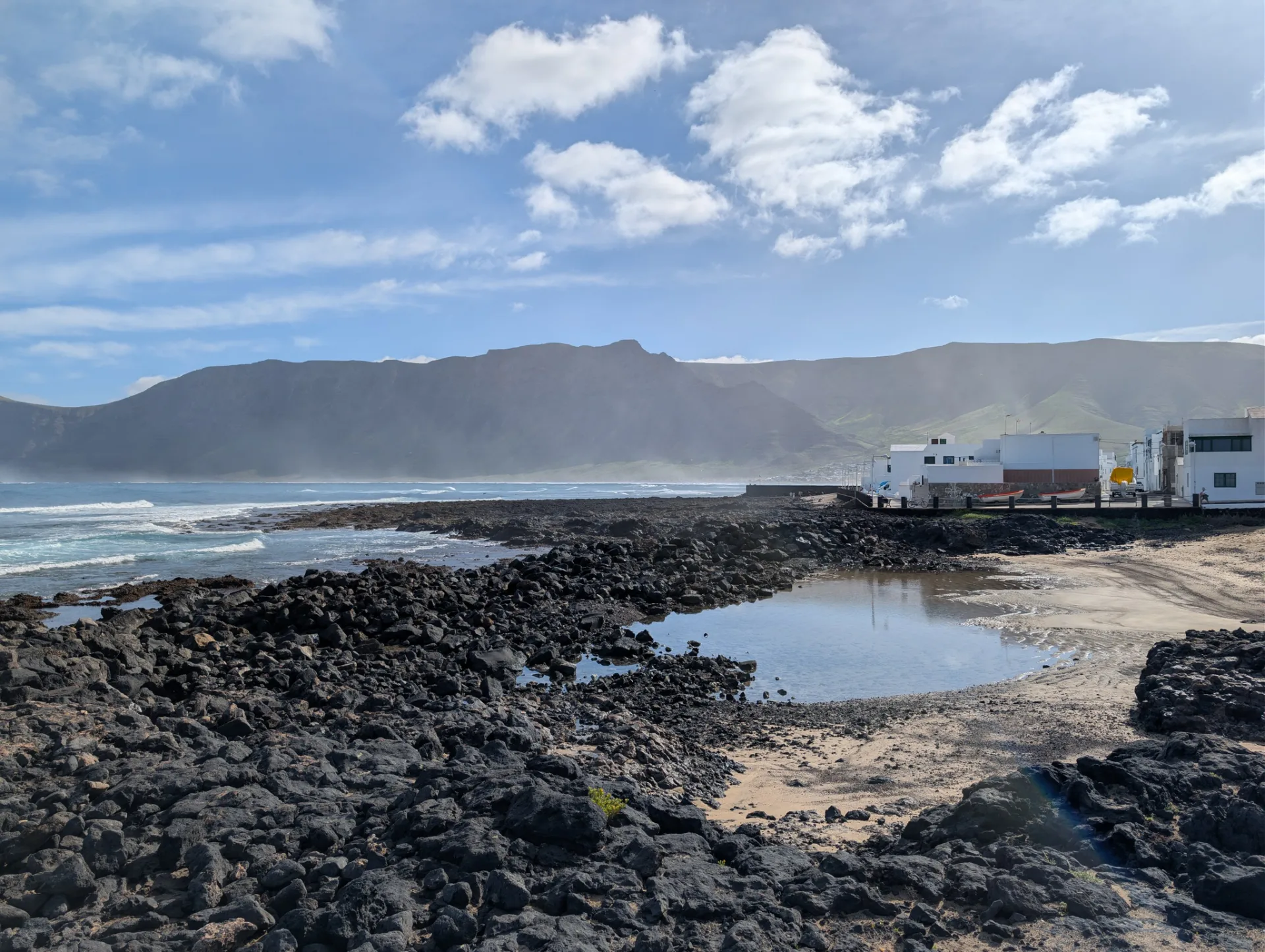 A small coastal settlement on the rocky shore, the Famara massif behind.